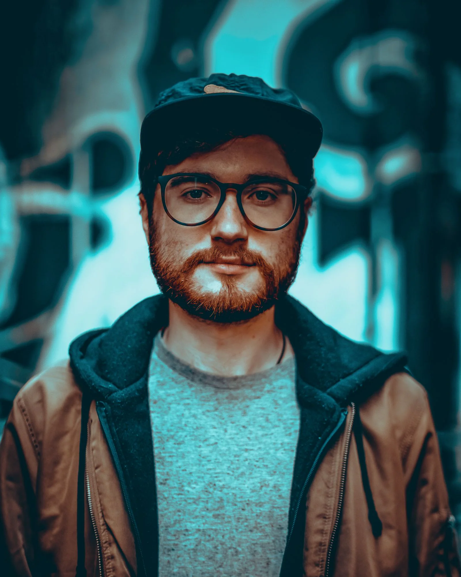 Scott Kehres in glasses, a cap and a tan jacket, in front of a teal graffiti wall